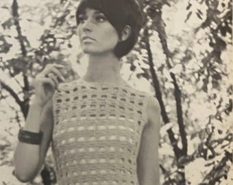 May include: A woman wearing a sleeveless crochet dress with a geometric pattern. The dress is white and has a square neckline. The woman has short, dark hair and is standing in front of a blurred background of trees.