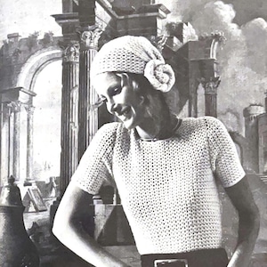 Può includere: Una donna indossa una maglia bianca a uncinetto a maniche corte e un cappello bianco a uncinetto. Il cappello ha un grande dettaglio floreale sul lato. La donna sorride e guarda di lato.