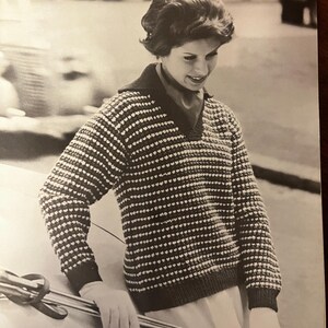 May include: A black and white striped sweater with a V-neck and a textured pattern. The sweater is worn by a person standing next to a car.