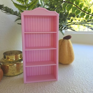 Puede incluir: Una pequeña estantería rosa con cuatro estantes. La estantería en miniatura tiene una parte superior decorativa y ranuras verticales en el panel posterior. Los estantes están vacíos. Un tarro dorado y una calabaza de fieltro están en el fondo.
