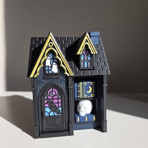 May include: A miniature black house structure featuring gold-trimmed gothic windows with white ghost silhouettes, a purple stained-glass door with a black raven, and an open side shelf displaying two blue books with gold moons and a white skull.