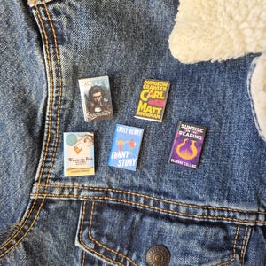 Op de afbeelding: Een spijkerjasje met vijf miniatuur boekenpinnen. De pinnen tonen boektitels zoals "Quicksilver", "Dungeon Crawler", "Winnie the Pooh", "Funny Story" en "Sunrise Reaping". De jas heeft een lichtblauwe wassing.