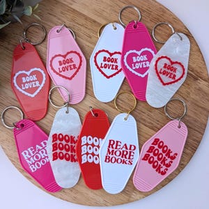 May include: A collection of motel-style keychains in various colors, including red, pink, and white. Each keychain features text such as "Book Lover" or "Read More Books" in a bold, red font. The keychains are arranged on a wooden surface.