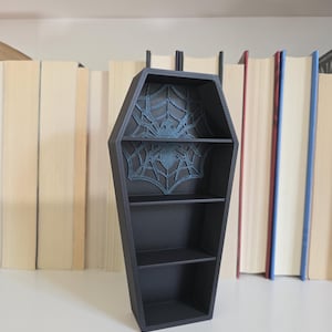 May include: A matte black coffin-shaped shelf unit featuring three horizontal tiers for storage. The back panel of the top section displays a raised, dark blue spider and web design, providing a textured focal point within the structure.