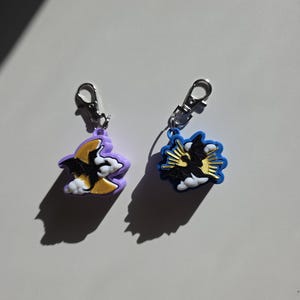 May include: Two rubber charms with silver-tone lobster clasps. The left charm is lavender and features a black bat flying in front of a yellow crescent moon and white clouds. The right charm is blue with a black bat, yellow sunburst, and white clouds.