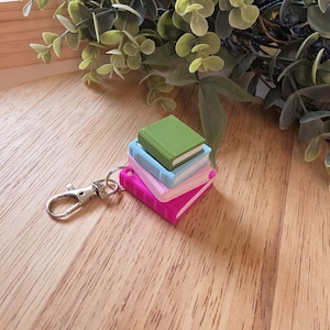 May include: A keychain featuring a stack of miniature books in pink, light blue, and green. The books have textured covers and are attached to a silver-colored clasp. The background includes greenery and a wooden surface.