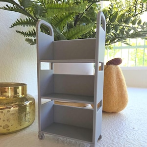 May include: A small, gray, three-tiered cart with wheels. The cart has a handle at the top and open sides. The cart is designed for storage and organization. The cart is approximately 12 inches tall.