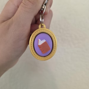 May include: A gold-colored oval keychain featuring a purple circular center with a raised, three-dimensional orange book icon. The keychain includes a silver-toned metal split ring and a lobster claw clasp for attachment to keys or bags.