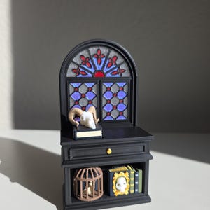 May include: A miniature black cabinet featuring an arched top with a blue and red stained glass window design. The cabinet includes a drawer with a gold knob, a top surface holding a book with a ram skull, and a lower shelf with a birdcage and books.