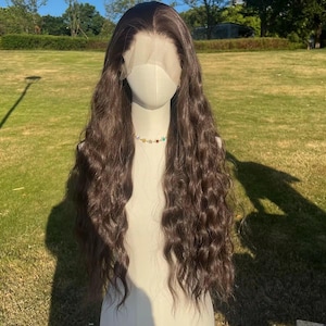 Puede incluir: Una larga peluca marrón ondulada con una parte delantera de encaje se muestra en un maniquí. La peluca presenta una parte natural y un patrón de ondas suaves y texturizadas. Un collar de cuentas de colores adorna el cuello del maniquí, añadiendo un toque de elegancia.