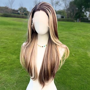 May include: A long, layered wig with brown, blonde, and light brown highlights. The wig features a natural-looking part and long, flowing strands. A colourful beaded necklace is around the mannequin's neck. The wig is displayed on a white mannequin head.