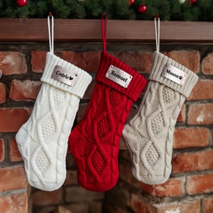 Peut inclure: Trois bas de Noël en tricot torsadé, blancs, rouges et beiges, suspendus à un manteau en bois. Chaque bas porte une plaque nominative en métal avec les noms "Cedric", "Samuel" et "Mason". Les bas sont suspendus devant une cheminée en briques.
