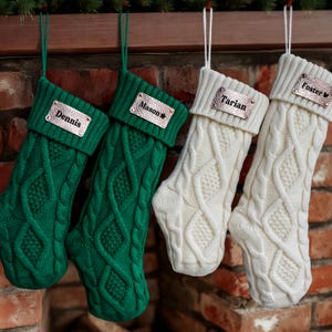 Peut inclure: Quatre bas de Noël en tricot torsadé, verts et crème, suspendus à une cheminée en bois. Chaque bas porte une étiquette nominative personnalisée en métal. Les noms sont Dennis, Mason, Tarian et Foster.