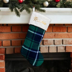 Peut inclure: Une chaussette de Noël avec un motif à carreaux vert et marine. Le haut de la chaussette est blanc avec le nom "Ronnie" brodé en or. La chaussette est suspendue à une cheminée blanche avec des décorations de Noël.