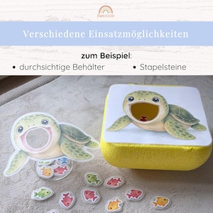 May include: A turtle-themed game set featuring two turtle cutouts with open mouths, a yellow foam container, and several small, colorful fish-shaped tokens in red, orange, green, and pink. The text reads: Verschiedene Einsatzm&ouml;glichkeiten zum Beispiel: durchsichtige Beh&auml;lter Stapelsteine.