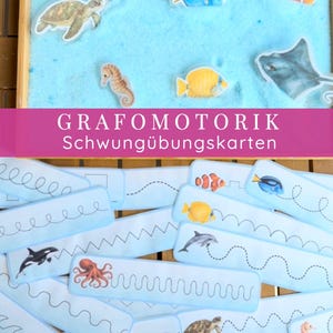 Könnte beinhalten: Ein Holztablett mit blauem Sand und Meeresbewohner-Ausschnitten, begleitet von hellblauen Papierstreifen mit gestrichelten Linien zum Nachspuren und Illustrationen von Meerestieren, mit dem Text GRAFOMOTORIK Schwungübungskarten.