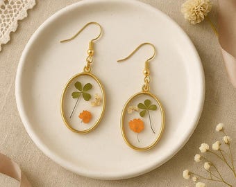 Puede incluir: Un par de pendientes ovalados dorados. Cada pendiente presenta un óvalo de resina transparente con un trébol de cuatro hojas, una pequeña flor naranja y diminutas flores blancas. Los pendientes se exhiben en un plato blanco.