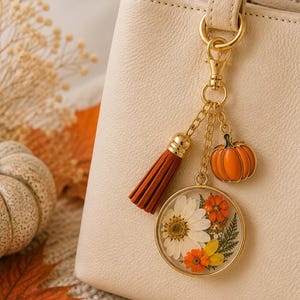 May include: A gold-toned metal bag charm attached to a cream-colored handbag. The charm features a round pendant containing pressed white and orange flowers, a small orange pumpkin charm, and a rust-colored fabric tassel on a gold chain.