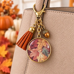 May include: A gold-toned metal bag charm featuring a circular pendant with a colorful autumn leaf print, a burnt orange suede tassel, and a small metallic acorn charm, all attached to a lobster clasp hanging from a beige textured handbag.