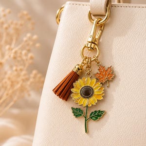 May include: A gold-toned metal bag charm attached to a cream-colored handbag. The charm features a yellow sunflower with a dark center, a green stem with leaves, an orange maple leaf, and a small brown tassel, all connected by a gold lobster clasp.