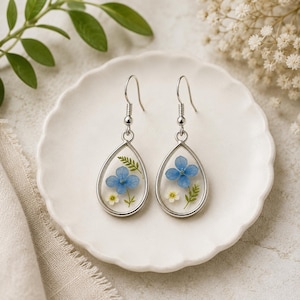 May include: A pair of silver-toned teardrop earrings featuring clear resin centers. Each earring contains pressed blue hydrangea petals, small white flowers, and green fern leaves, suspended from a simple silver-toned metal hook wire.