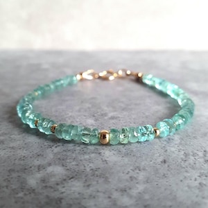 Puede incluir: Una delicada pulsera con pequeñas cuentas de piedras preciosas de color verde azulado claro ensartadas. La pulsera está adornada con cuentas de color dorado y un cierre de color dorado. La joya se exhibe sobre una superficie gris.