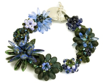 Op de afbeelding: Een kralenarmband met een zilveren sluiting, bestaande uit blauwe en groene glazen kralen die in een bloemmotief zijn gerangschikt.