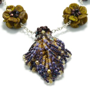 Op de afbeelding: Een kralenketting met een kwast hanger. De ketting heeft bruine en gouden bloemvormige kralen en een zilveren ketting. De kwast hanger is gemaakt van paarse en gouden kralen.