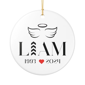 Puede incluir: Adorno navideño redondo de cerámica blanca con una cuerda dorada. Presenta el nombre "LIAM" en negro, con alas de ángel y un halo encima. Debajo, los años 1993 y 2024, separados por un corazón rojo.