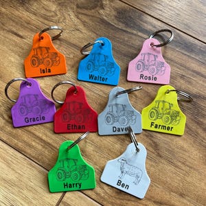 May include: A selection of brightly coloured keyrings, each with a tractor design and a name. The keyrings are in shades of orange, blue, pink, purple, red, grey, yellow, green, and white. Names include Isla, Walter, Rosie, Gracie, Ethan, Dave, Farmer, Harry, and Ben.