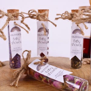 May include: A set of small glass bottles filled with dried flowers and herbs, each with a tag that reads "Oliver's Baby Shower" and the date "20.05.2021". The bottles are tied with twine and have a rainbow design on the tags.