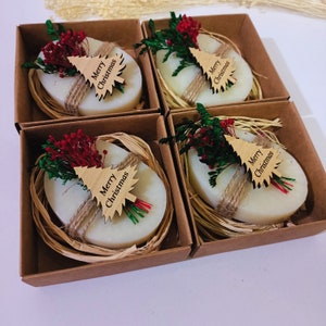 Peut inclure: Quatre petites boîtes cadeaux avec des couvercles bruns et des savons ovales blancs à l'intérieur. Chaque savon est décoré d'un petit sapin de Noël en bois avec les mots "Merry Christmas" dessus. Les boîtes sont remplies de matière ressemblant à de la paille et décorées de petites branches de feuillage rouge et vert.