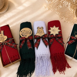May include: Five rolled scarves in red, dark green, and white, each featuring a plaid ribbon and a wooden ornament engraved with Merry Christmas. The scarves have fringed ends and are presented in cardboard boxes or tied with twine.