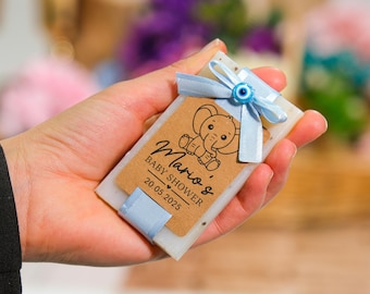 May include: A blue and white baby shower favor soap with a brown tag that says "Mario's Baby Shower 20.05.2025" and features a cartoon elephant. The tag is tied with a blue ribbon and a blue evil eye charm.