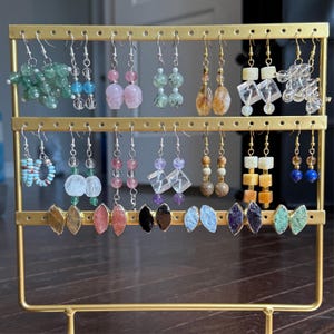 May include: A gold earring display stand showcasing multiple pairs of earrings. The earrings feature diverse gemstones and beads in shades of green, pink, clear, brown, and blue. The earrings are presented on a gold-coloured stand.