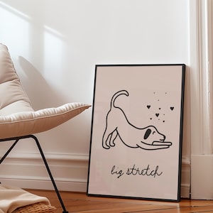 May include: Black framed art print featuring a minimalist line drawing of a dog stretching, with hearts and the words "big stretch" in a script font. The artwork has a neutral color palette and is leaning against a white wall.