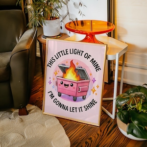 Peut inclure: Une affiche encadrée avec une illustration d'une benne à ordures rose souriante avec une flamme. Le texte dit : « This little light of mine, I'm gonna let it shine ». L'œuvre est dans un cadre doré sur fond rose.