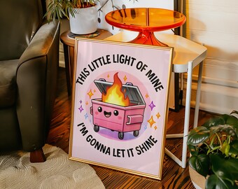 May include: A framed print featuring a pink dumpster with a smiling face and a flame. The text around the dumpster reads "This little light of mine, I'm gonna let it shine." The artwork has a gold frame and is set against a pink background.