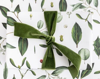 May include: A wrapped gift with white botanical print paper featuring green leaves and sprigs. A green velvet ribbon is tied in a bow around the gift. The gift wrap design includes illustrations of various green leaves and small red berries.