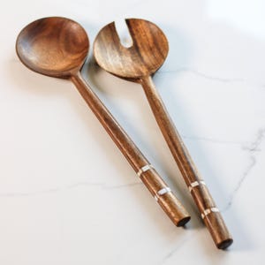 May include: Two wooden salad servers with a rich brown finish. One is a spoon, the other a slotted fork. The handles have decorative white inlays. The servers are designed for serving salads and other dishes.