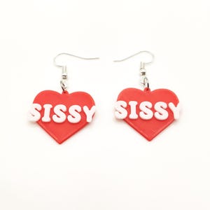 May include: A pair of red heart-shaped earrings with the word "SISSY" in white lettering.