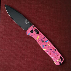 Könnte beinhalten: Ein pinkfarbenes Klappmesser mit schwarzer Klinge und einem Griff, der mit bunten Streuseln verziert ist. Die Klinge hat ein Schmetterlingslogo. Das Messer ist geöffnet und angewinkelt, vor rotem Hintergrund.