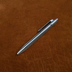May include: A silver metal pen with a clip, lying diagonally on a brown leather surface. The pen has a sleek, modern design with a shiny finish. The pen's tip is pointed, and the clip is attached to the pen's body.