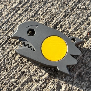 Può includere: Un utensile multifunzione in metallo grigio opaco a forma di testa di squalo stilizzata con un inserto circolare giallo brillante al centro. L'utensile presenta un'apertura esagonale, un bordo seghettato simile a denti e un foro sul retro.