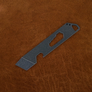 May include: A dark gray multi-tool with a blade, bottle opener, and other features. The tool is rectangular with a key ring hole and various cutouts. The blade has a beveled edge. The tool is shown on a brown leather surface.