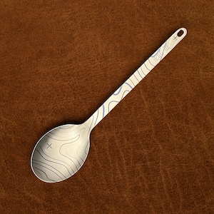 May include: A silver-toned metal spoon featuring a laser-etched topographic map pattern across the bowl and handle. The handle includes a small circular hole at the end and a rectangular mark with the text "TITANIUM" near the top.