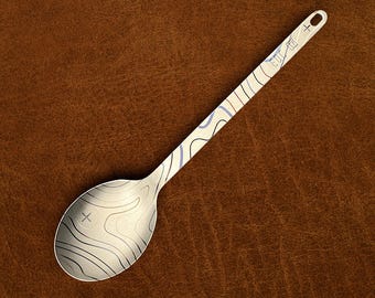 May include: A silver-toned metal spoon featuring a laser-etched topographic map pattern across the bowl and handle. The handle includes a small circular hole at the end and a rectangular mark with the text "TITANIUM" near the top.