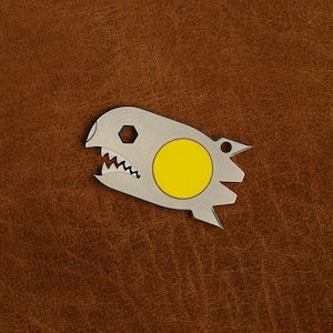 May include: A silver-toned metal multi-tool shaped like a stylized shark head with a bright yellow circular inlay. The tool features a hexagonal cutout, a serrated edge resembling teeth, and a small circular hole at the rear for attachment.