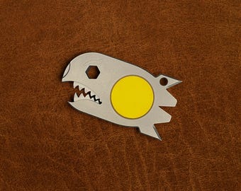 May include: A silver-toned metal multi-tool shaped like a stylized shark head with a bright yellow circular inlay. The tool features a hexagonal cutout, a serrated edge resembling teeth, and a small circular hole at the rear for attachment.