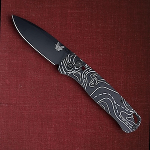 May include: Black folding knife with a topographic map design on the handle. The blade is black with a butterfly logo. The handle is black with a white topographic pattern. The knife is on a red background.